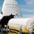 Looking like the mouth of an enormous great white shark, a white and blue Russian cargo plane is opened to reveal the large Centaur upper stage engine that will blast the Mars Reconnaissance Orbiter to Mars.  The large cylindrical engine is covered in a protective white tarp and is being carefully marshaled off the plane and down a bright yellow ramp by a team of technicians.  The bright blue sky, streamed with wispy clouds is in the background along with a line of brilliant green trees.