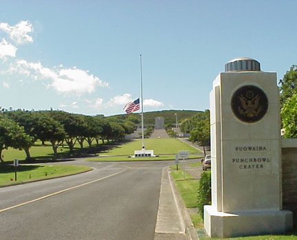 A picture of the entrance gate and road leading to the Punchbowl-shaped cemetery. A flagpole stands midway flying the American flag at half mass.