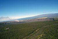 Aerial view looking east toward across Kīlauea's summit caldera toward the plume from Halema'uma'u. The summit and enormous southwest flank of Mauna Loa is in the background.