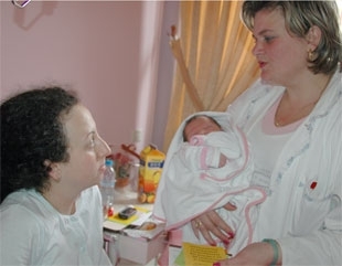 A woman in a white medical coat holds an infant while speaking to a young woman