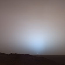 This color panorama shows the dark silhouette of a rugged cliff extending downward from left to right and beneath a flat horizon on the right. In the distance, the yellowish orb of the sun is beginning to sink beneath the hills that form the rim of Gusev Crater. All around the sun, glowing light streams upward and outward until it disappears into the purplish darkness of twilight.