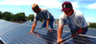 Photo of two young men installing a solar panel in a row of solar panels; another row of solar panels can be seen in the background.