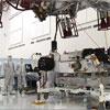 These three images show the progression of 'stacking' the Mars Science Laboratory rover and its descent stage in one of the Jet Propulsion Laboratory’s 'clean room.' In this image, the car-size rover is in the middle of the picture with several team members surrounding it.  The team members are all dressed in special head-to-toe white suits, called 'bunny suits.'  One team member is holding on to a tether to guide the large insect-like descent stage down on top of the rover.  The descent stage looms high in this image.