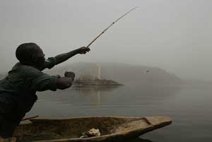 A man fishes in a dugout canoe on the Obangui river, with the city of Bangui in the background, Central African Republic. March 9, 2004. [© AP Images]