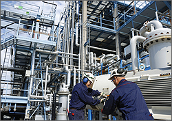 Photo of two men in blue coveralls and white hard hats working together at a petroleum refinery. The men are facing each other and one man has his back to the camera. CREDIT:  Lagereek at iStock.com.