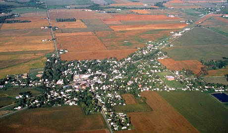 Panoramic view of a small town with a field in the foreground and mountains in the background.  The steeple of a church is visible above the roofs and the tops of the trees.