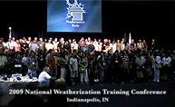 Photograph of the attendees of the National Weatherization Training Conference, lined up in four long rows on a stage.  Beneath them, the text reads, '2009 National Weatherization Training Conference, Indianapolis, TN.'  They stand in front of a large blue screen with the Weatherization Program's logo on it-a house surrounded by the wind, rain, sun, and snow, above the words 'Weatherization Works.'