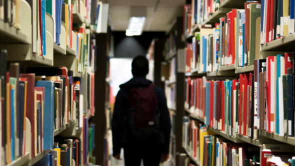 A photograph of the back of a student carrying a backpack as they walk down an aisle of books in a library.