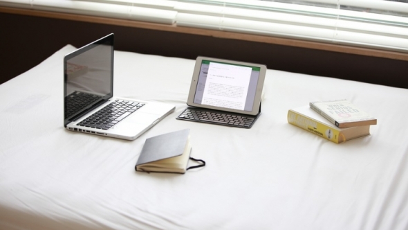 A photograph of a laptop, tablet computer, notebook, and two books on a bed. 