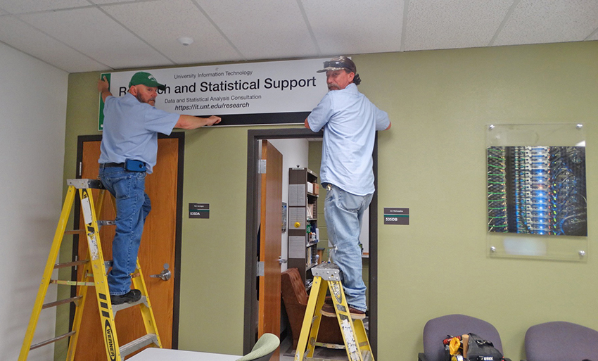 Brent Lewis and Mike Chenault, Facilities Services, hang the new R&SS sign