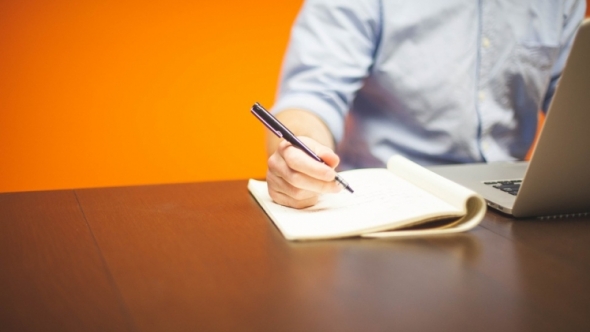 A photo of a person sitting at a desk with an open laptop and open notepad.  