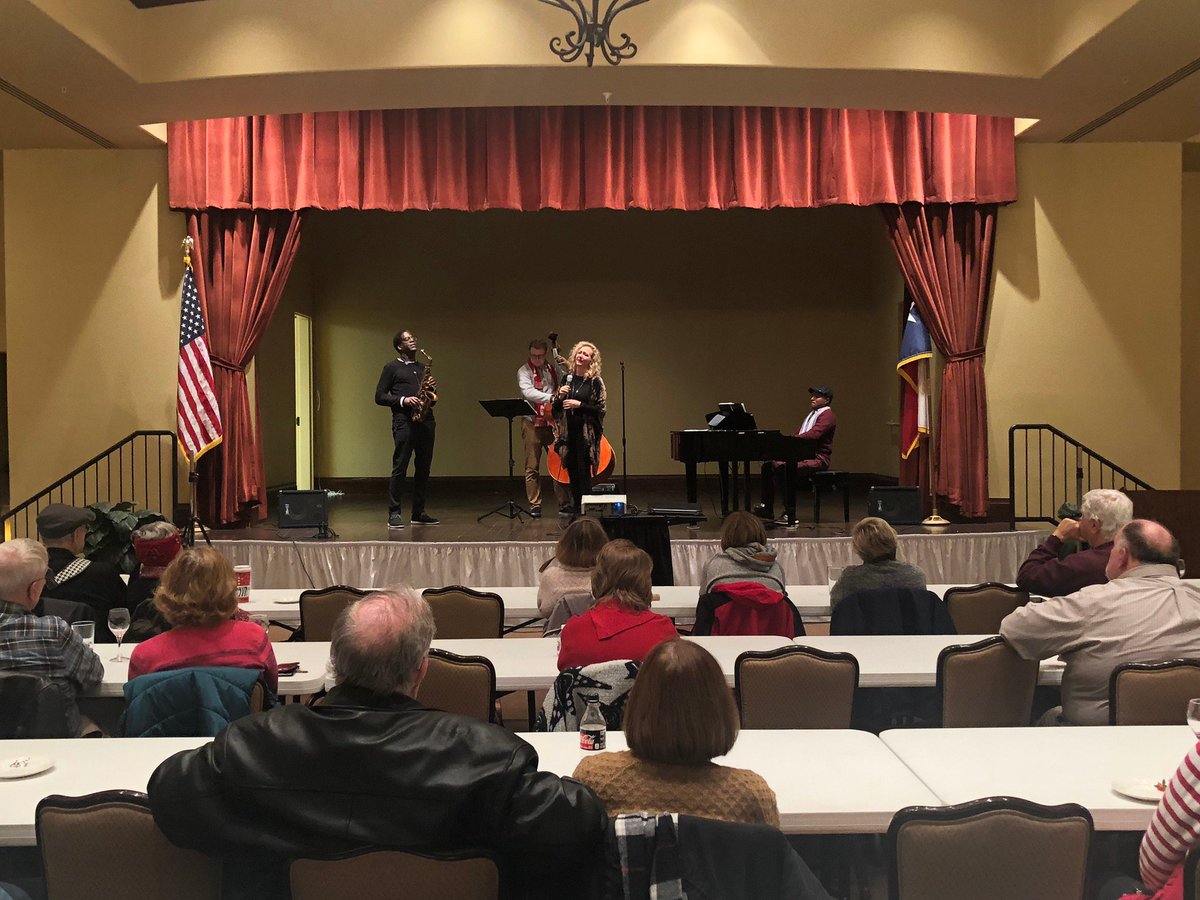 A ballroom full of senior adults enjoy wine, cheese, and crackers. On stage at the front of the room, Brad Leali plays saxophone, Carla Helmbrecht sings, and they are accompanied by a piano player and an upright bass player.