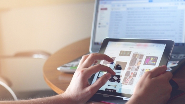 A photo of a person using an iPad in the foreground with a desktop computer in the background. 