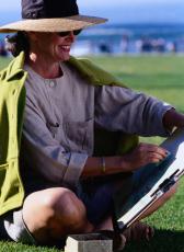 Photograph of a woman in a hat and sunglasses drawing in a park