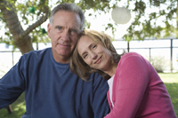 A middle-aged man and woman sitting.  Both are smiling into the camera.  The woman is leaning onto the man with her head on his shoulder.