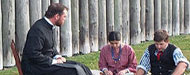 A man sitting on a chair and two younger kids sitting in the grass talking.