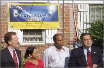 Photo of two men and a husband and wife standing in front of microphones set up in front of their home.