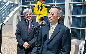 Photo of a man in a business suit standing at a microphone answering media questions while another man in a business suit listens.