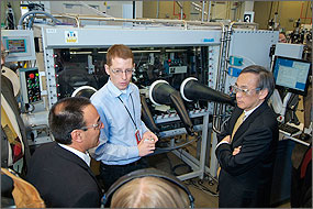 In photo, three men wearing safety glasses stand in front of a large glass box containing scientific equipment. Protruding from the front of the box are large black rubber gloves used to work with the equipment inside.