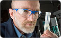 Photo of a man with a trim white beard wearing safety glasses and a dark blue lab coat, looking closely at a test tube containing a clear liquid.