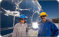 Photo of a young woman and a man with a moustache, both wearing hardhats and dark safety glasses, standing outside in front of a large curved metal reflector. The man is holding a small square-shaped sample of the silver reflective surface, which replaces heavy glass mirrors in the design. The large reflector is streaked with snow and frost after a recent winter storm.