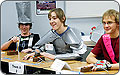 Photo of three young men sitting at a table with their fingers on buttons controlling a buzzer. The teammates are costumed in a stovepipe hat, left, and armor, center, made from duct tape. The rightmost has a bright red banner across his chest.