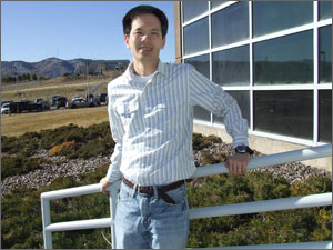 Photo of a young man leaning on a railing in front of a brown building. Mountains are in the background.