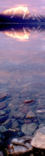 Sunset over crystal clear Lake McDonald in winter showing rocks on the lake bottom and snowcapped mountains in the distance.