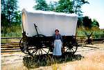 Woman in pioneer dress stands next to replica covered wagon