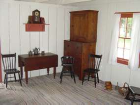 Corner of an interior room with white wooden walls and wooden furniture: two chairs, a high chair, a tall cupboard, and a shelf with books and a clock.