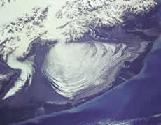 The massive lobe of Malaspina Glacier in Alaska is clearly visible in this photograph taken from a Space Shuttle flight in 1989.  Agassiz Glacier is the smaller glacier to the left.  The Malaspina Glacier is one of the most famous examples of this type of glacier, and is the largest piedmont glacier in the world.  Spilling out of the Seward Ice Field (visible near the top of the photograph), it covers over 5,000 square kilometers as it spreads across the coastal plain. (Photo courtesy of SPACE.com and NASA.)