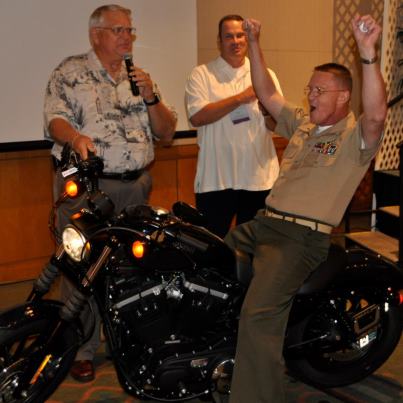 Photo: Marine Corps. Chief Warrant Officer Anthony John Kokesh shouts for joy as he sits on the Harley-Davidson 883 Sportster he won Aug. 23, in a contest, sponsored by Dr. Pepper Snapple Group. Looking on is Bob Schneller (left) and Dean Purcell, executives with Dr. Pepper Snapple Group. CWO5 Kokesh, a chemical biological, radiological and nuclear defense officer assigned to G-34 Marine Forces, Pacific, Hawaii, entered the contest at the Schofield Barracks Commissary and was one of four finalists – one from each commissary on Hawaii – who drew motorcycle keys at the Hawaii Prince Hotel, Honolulu. Only the key CWO5 Kokesh drew started the engine. The three other finalists, Dan Shishido, Honolulu; Katie Stahl, Kaneohe, Hawaii and Nicole Harbin, Ewa Beach, Hawaii, won 32” high-definition TVs.