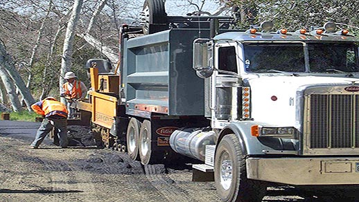 Workmen compress asphalt alongside of a truck and asphalt paver.