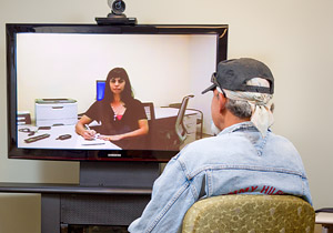 Veteran sitting, looking at a large TV screen where a mental health care worker is pictured, ready to take notes