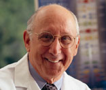 Image of caucasian man, wearing glasses, a light purple shirt, dark purple tie, and a white lab coat, sittling in a lab