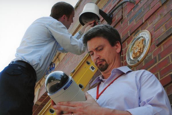 April 18, 2004:  A DS security technical specialist (foreground) inspects a surveillance camera while his colleague works on the mounting hardware at a United States diplomatic facility.  (Source: DS Records)