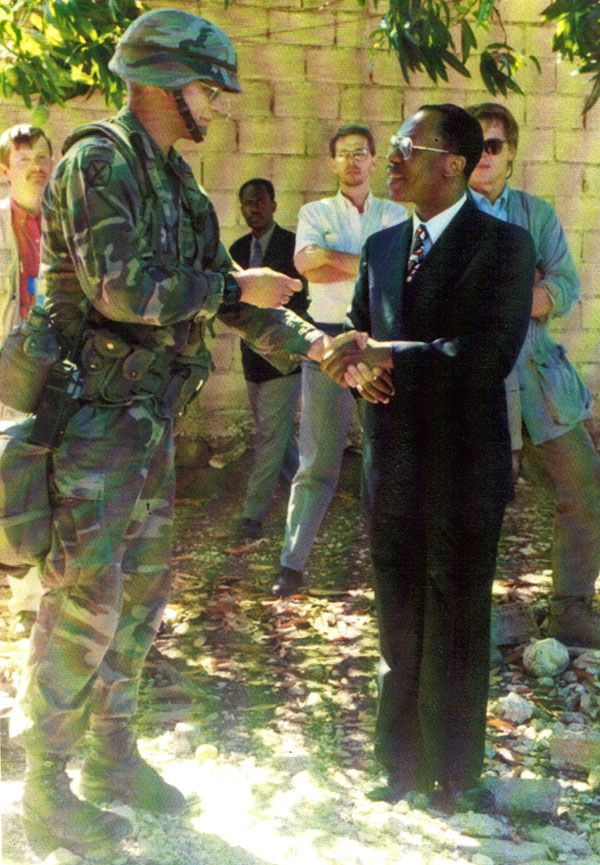 January 1995:  DS protective personnel (left, center in white shirt, and at right) look on as a uniformed U.S. Army battalion commander with the 10th Mountain Division presents Haitian President Jean-Bertrand Aristide with a souvenir battalion coin.  (Source: Private Collection)