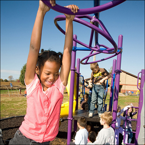 A little girl happily swings on the playground monkey bars while several other children play in the background.