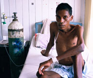 Emaciated man sits up in bed, shirtless, next to large tank with tubes in nose