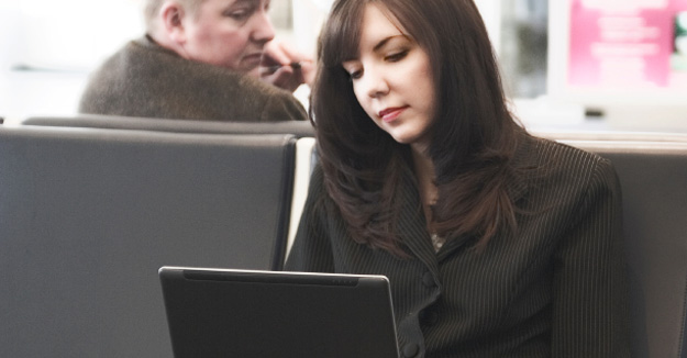 Woman on her laptop at an airport, unaware that a man is looking over her shoulder at the screen