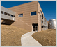 Photo of a walkway leading up a small incline to the entrance door of a two-story brick building with a silver tower on the right-hand side and a silver breezeway entering at the second floor on the left.