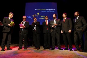 Photo of a group of men standing in a spotlight beneath a projected sign that reads “EU Sustainable Energy Week Awards Ceremony 2011.”  