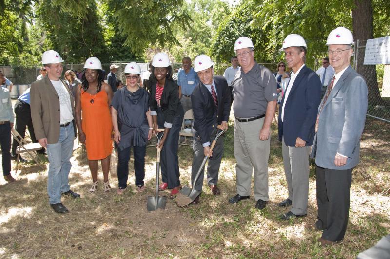 Photo of a group of people wearing hardhats. In the middle, two people hold shovels of dirt.