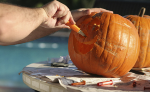 Adult carving smiley face pumpkin with a halloween carving tool. Pumpkin is on a flat table.