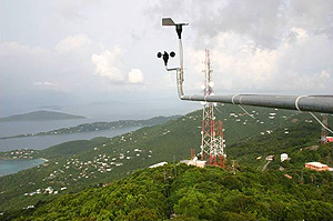 Photo of a wind anemometer installed on a mountaintop overlooking an island landscape. The ocean can be seen in the distance.