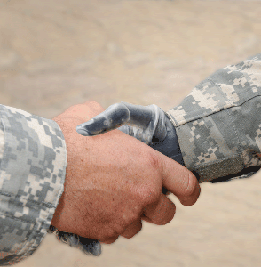 Cover Photo : A close-up of a handshake between two service members in camouflage uniforms. One person’s hand is a prosthesis. 
