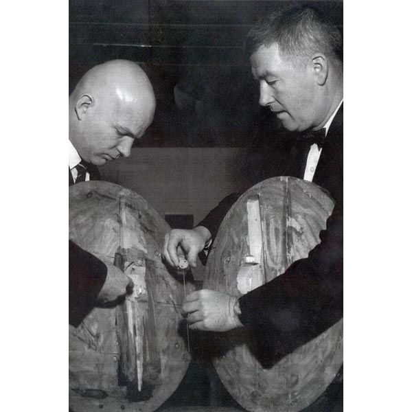 Circa 1961:  Director of Security John Reilly (right) holds the cavity resonator 'bug' microphone found inside a carved wooden image of the United States Great Seal, presented by Soviet officials to the U.S. Ambassador to the Soviet Union in 1948.  The DS agent at left points to where the bug was placed in the carving. (Source: U.S. Department of State Archives) 
