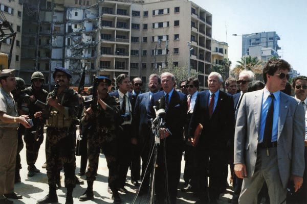 April 28, 1983:  U.S. Secretary of State George Shultz (at microphone) is surrounded by Office of Security special agents at the recently bombed U.S. Embassy in Beirut, Lebanon.  U.S. Ambassador to Lebanon Robert Dillon, with leather case, stands beside Secretary Shultz. (Source: Associated Press)