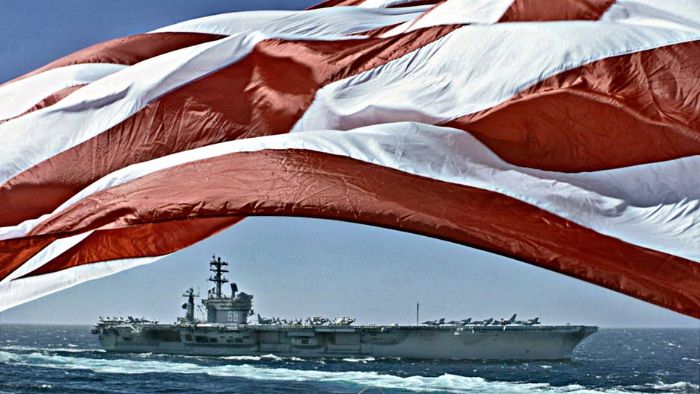 An aircraft carrier steams along as the American Flag waves proudly in the wind. 