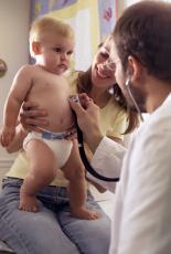 Fotografía de un doctor usando un estetoscopio para oír el latido de corazón de un bebé mientras la mamá lo sostiene